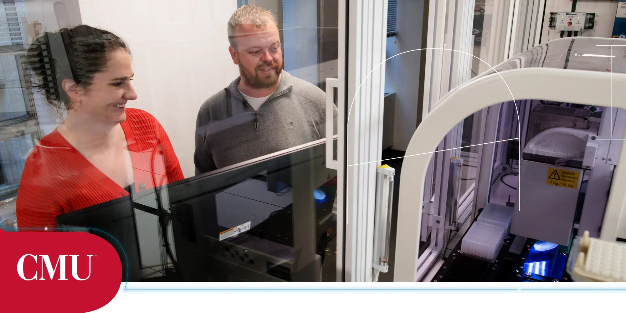 Two Carnegie Mellon University scientists looking at automated lab equipment, behind glass.